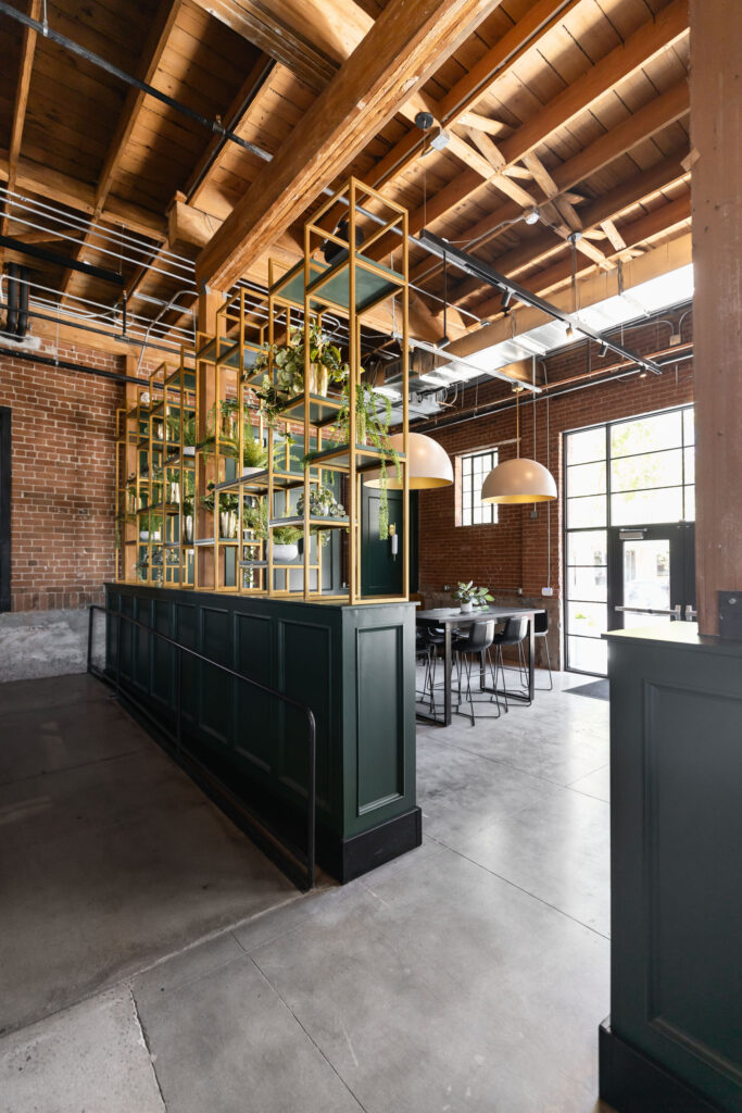 Contemporary restaurant space with dark green paneling, brass plant dividers, dome pendant lights, and brick walls.