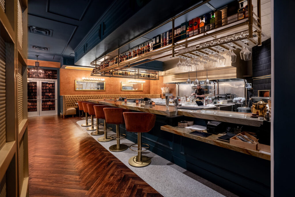 Upscale bar with herringbone wood floors, cognac leather barstools, tufted banquette, and suspended glassware rack above stainless counter.