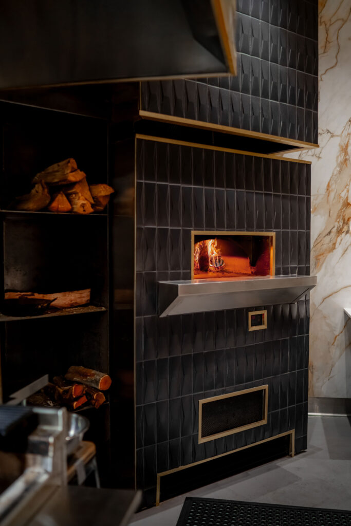 Modern tiled pizza oven with glowing fire, brass accents, stacked firewood, and marble wall detail.