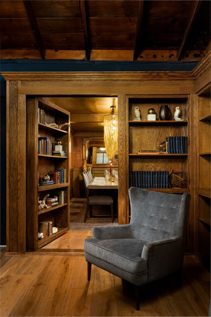 Intimate library nook with wooden built-in bookshelves, hidden doorway, gray accent chair, and warm pendant lighting.