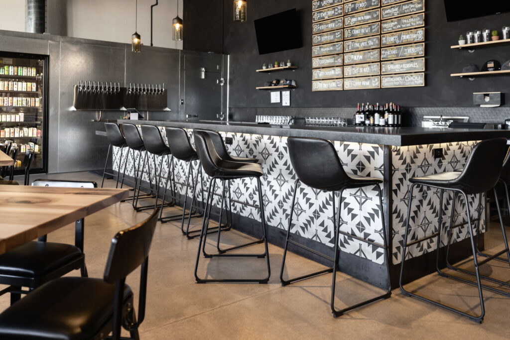 Modern bar featuring patterned tile front, black leather barstools, wooden tables, and beer taps along dark walls.