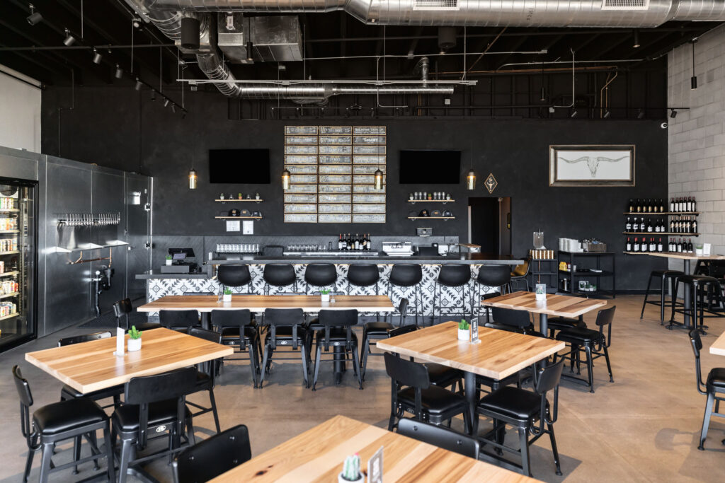 Spacious taproom with black ceiling, wooden dining tables, exposed utilities, and black bar with geometric tile accents.